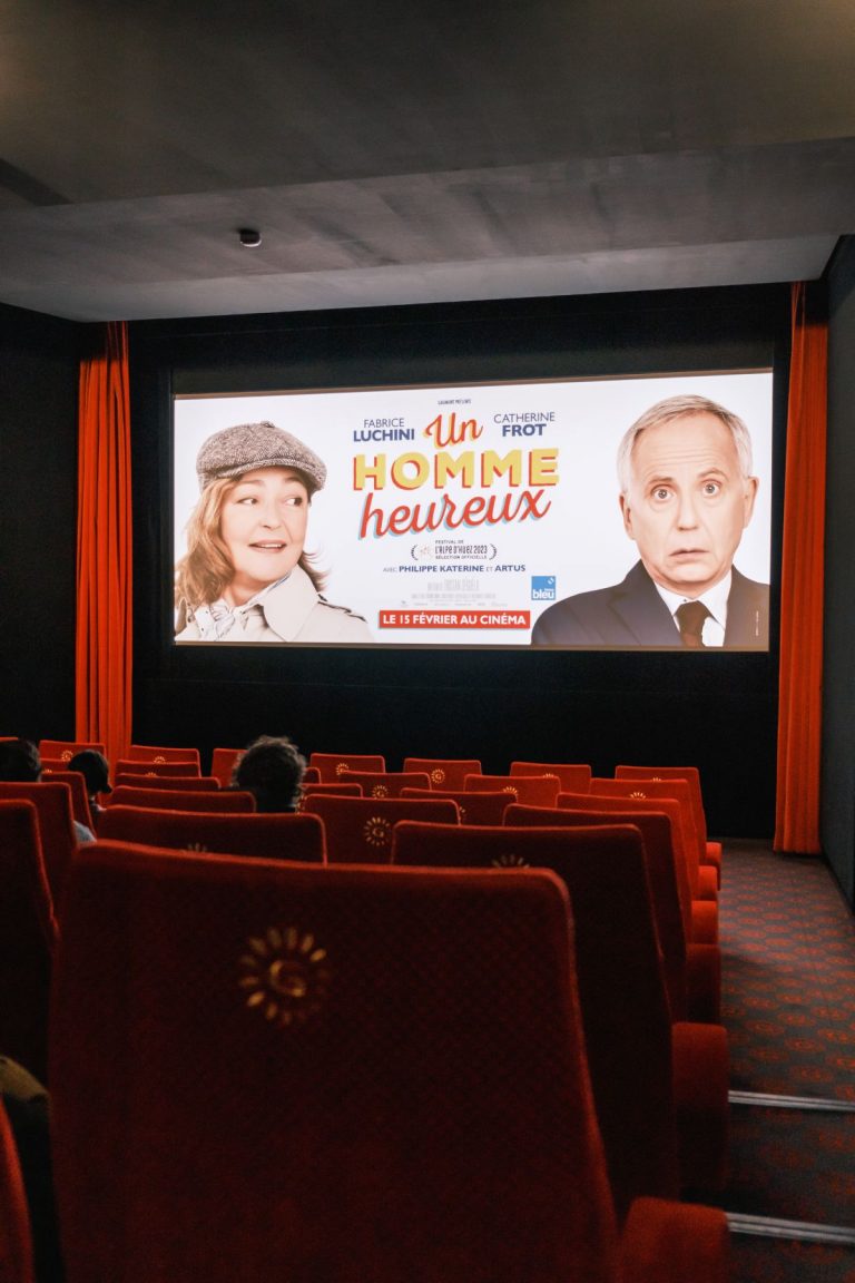 Salle de cinéma prise du fond de la salle, les sièges sont vides. Sur l'écran est projeté l'affiche d'Un Homme Heureux avec les portraits de Catherine Frot souriante et Fabrice Luchini circonspect.