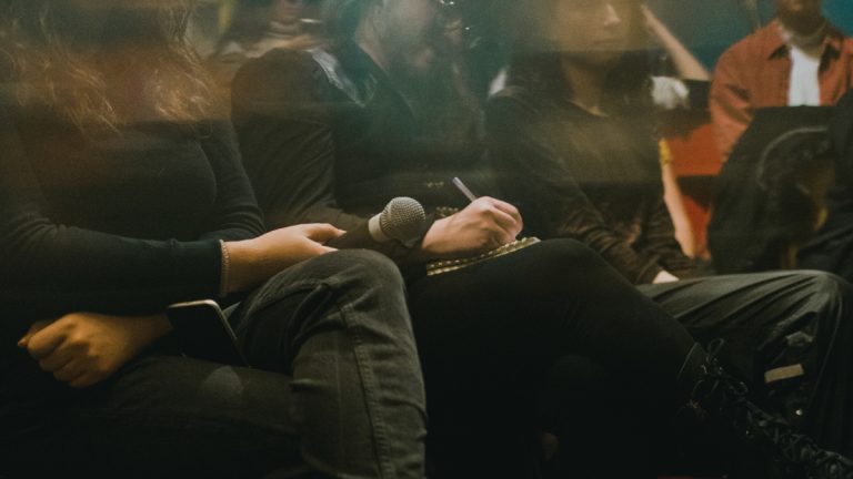 Photo du public, seuls les bustes de personnes assises sont visibles, une main d'une personne tient un micro, une autre personne écrit sur un papier poser sur un genou.